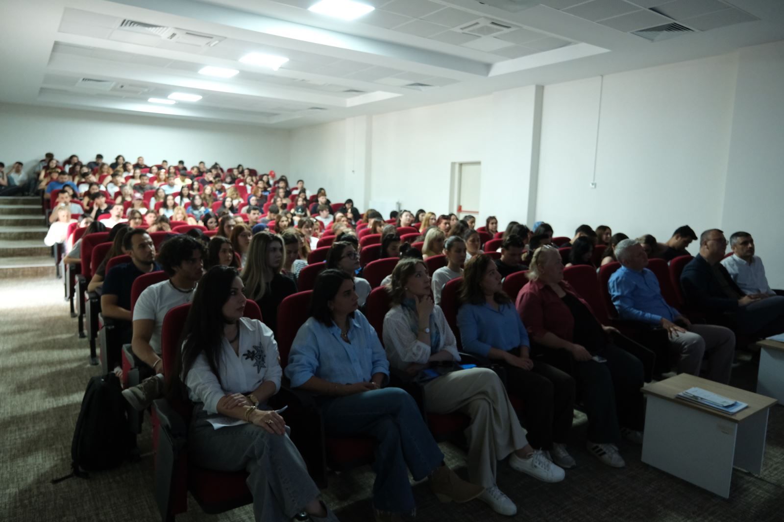 In the conference hall, students and participants are seated on red chairs, attentively following the presentation.